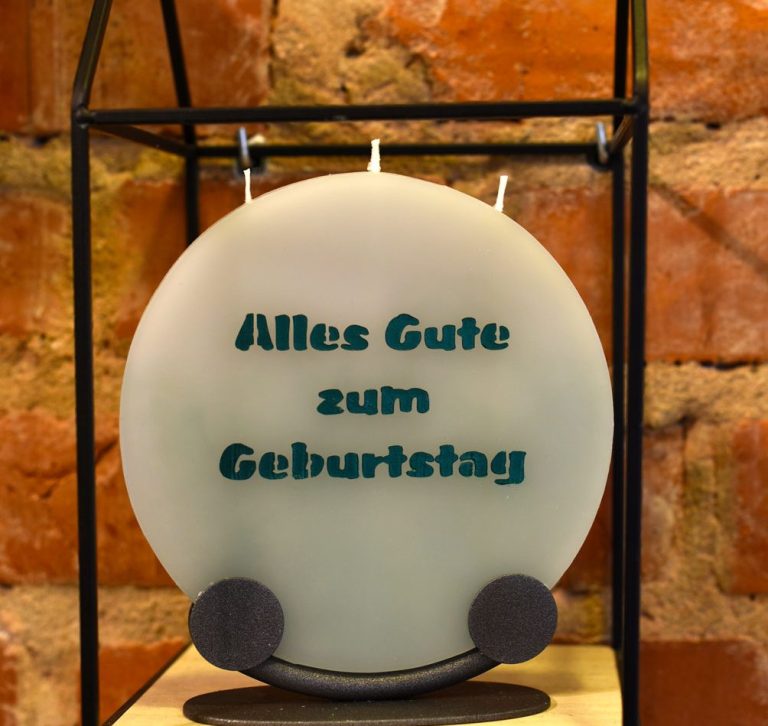 Runde Kerze mit der Aufschrift 'Alles Gute zum Geburtstag' vor einer Backsteinwand